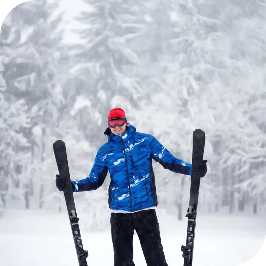 A man in winter clothing holding skis in front of a wintry forest.